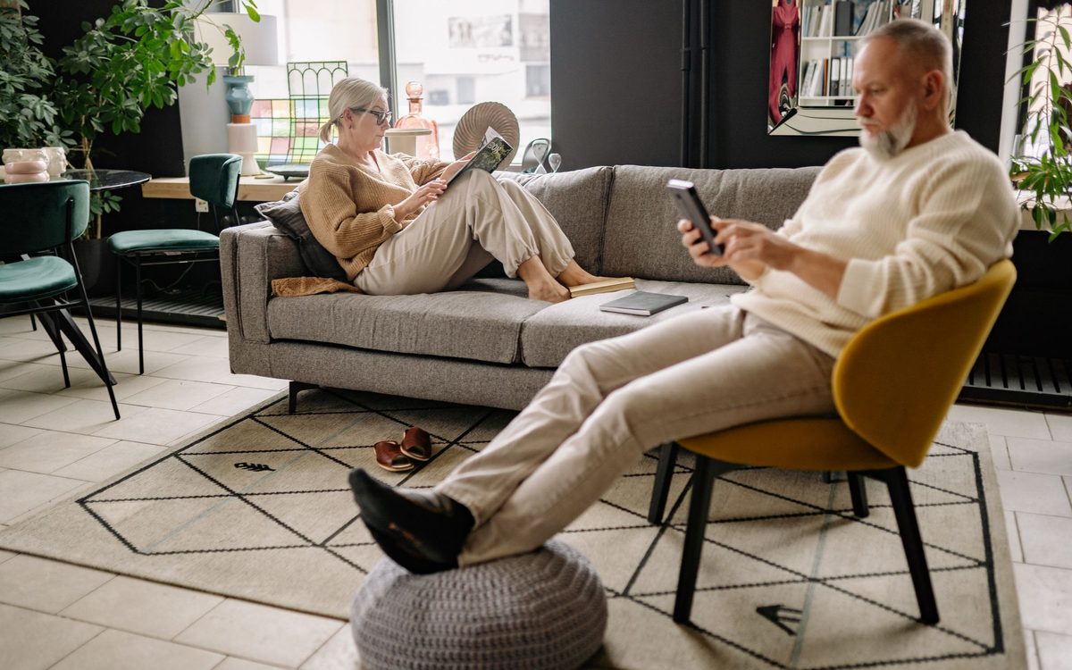 Older man resting in a chair with his feet up on a pouf while a woman reads on the sofa, a relaxing recliner-style setup for seniors.