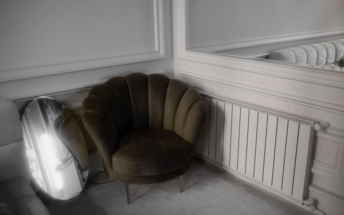 Dark olive velvet scalloped-back armchair in the corner of a paneled room beside a tilted oval mirror and a white radiator