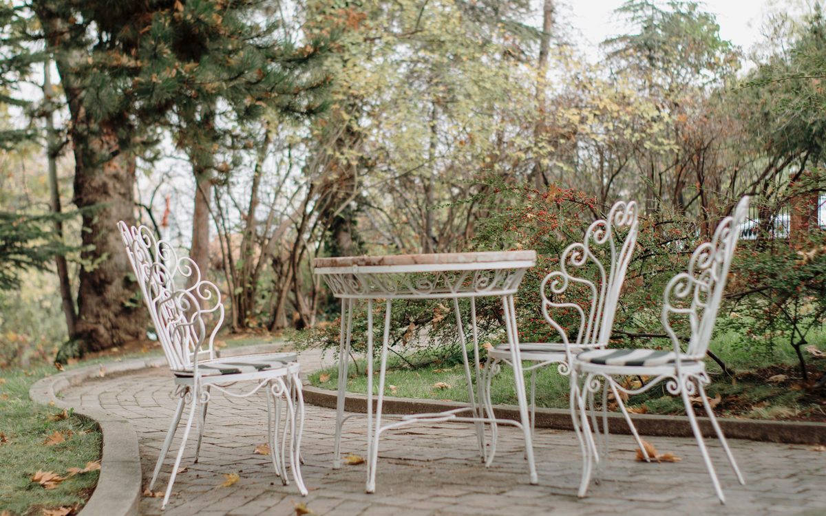 Ornate white metal chairs and a round table on a brick patio path surrounded by trees