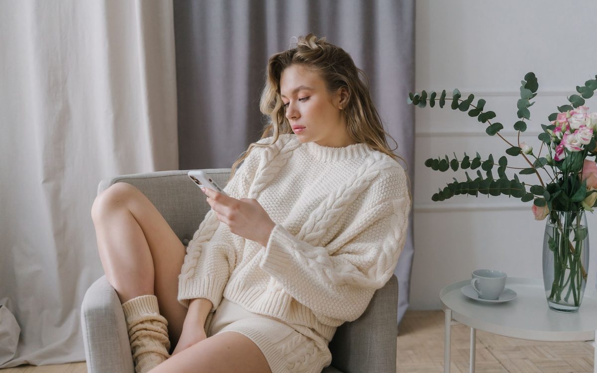Woman in a cozy sweater using a smartphone while sitting in a padded gray armchair
