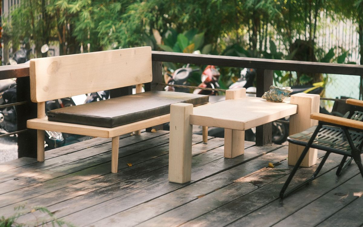 Pale wooden outdoor furniture, a cushioned bench and low table, on a shaded wood deck among green plants.