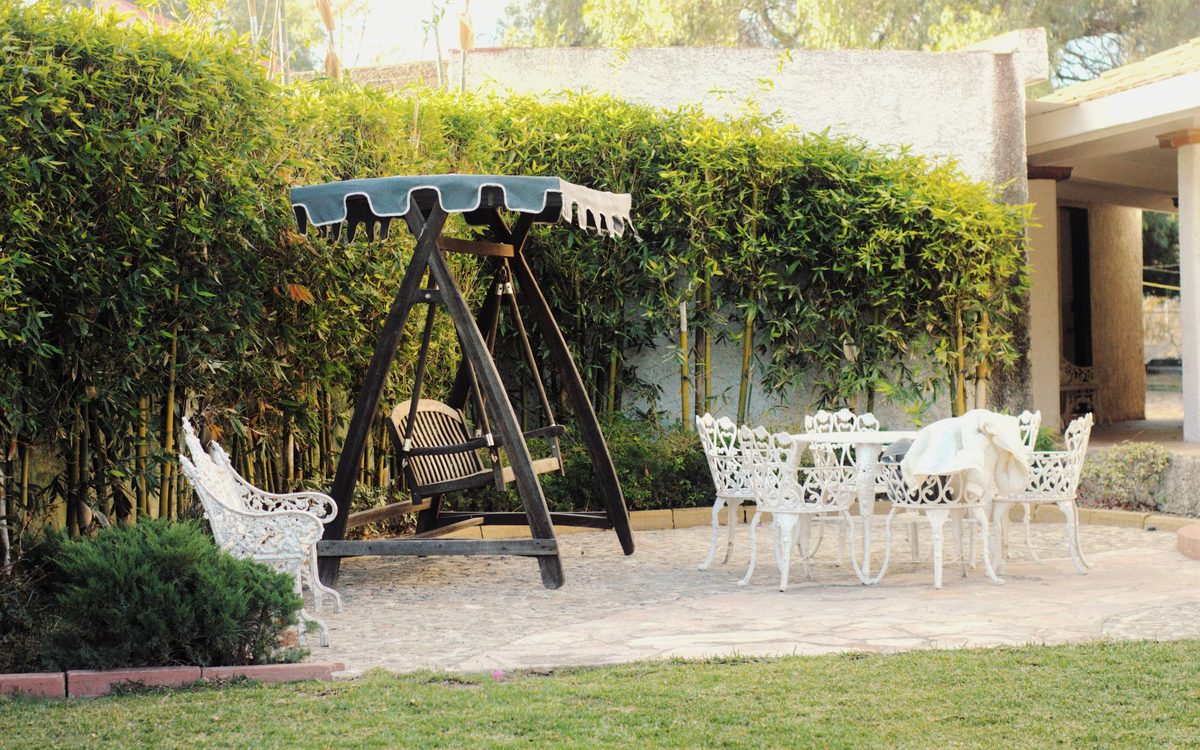 White ornate cast metal chairs and a round table on a stone patio, with a wooden canopy swing in front of a leafy hedge.